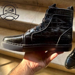 May include: Black high-top leather sneaker with a textured, crocodile-like pattern. The shoe features a lace-up design, a black sole, and a logo that says "RIGELLI LEATHER HAND CRAFTED".