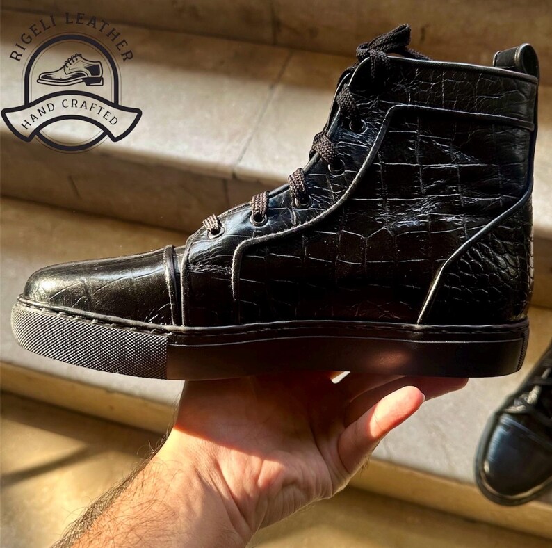 May include: Black high-top leather sneaker with a textured, crocodile-like pattern. The shoe features a lace-up design, a black sole, and a logo that says "RIGELLI LEATHER HAND CRAFTED".