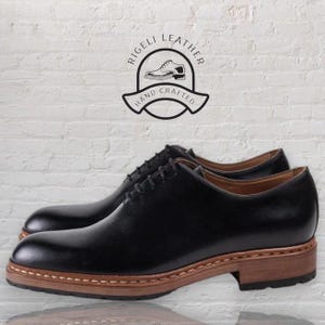 May include: A pair of black leather lace-up shoes with a brown leather sole. The shoes have a classic design and are made with high-quality materials. The shoes are hand-crafted and feature the text "Rigelli Leather Hand Crafted".
