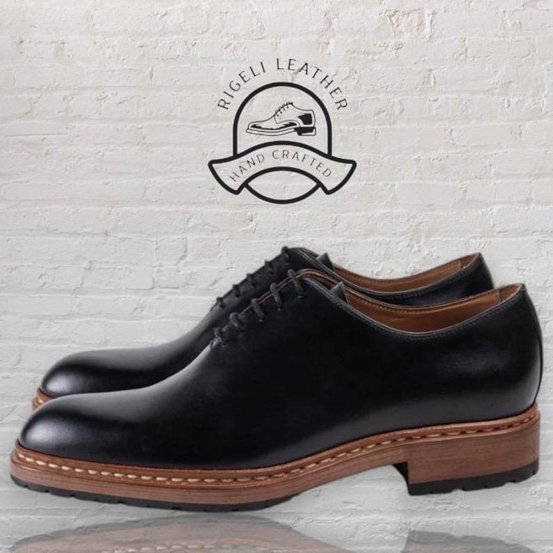 May include: A pair of black leather lace-up shoes with a brown leather sole. The shoes have a classic design and are made with high-quality materials. The shoes are hand-crafted and feature the text "Rigelli Leather Hand Crafted".