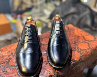 Könnte beinhalten: Ein Paar schwarze Oxford-Schuhe aus Leder mit Zehenkappe, geschlossenem Schnürsystem mit dünnen schwarzen Schnürsenkeln und polierter Oberfläche. Die Schuhe sind mit einer abgerundeten Zehenpartie und einem sichtbaren Absatz gestaltet und zeigen eine schlanke Silhouette.