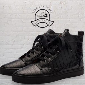 May include: Black high-top sneakers with a textured, crocodile-like pattern. The sneakers have black laces and soles. The image includes a logo that says "Rigeli Leather Hand Crafted".