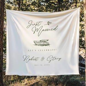 May include: A white fabric banner with the words "Just Married" and a heart symbol in elegant script. Below is a drawing of a boat, followed by "LET'S CELEBRATE!" and the names "Robert & Stacy" with a date. The banner is hanging between two trees.