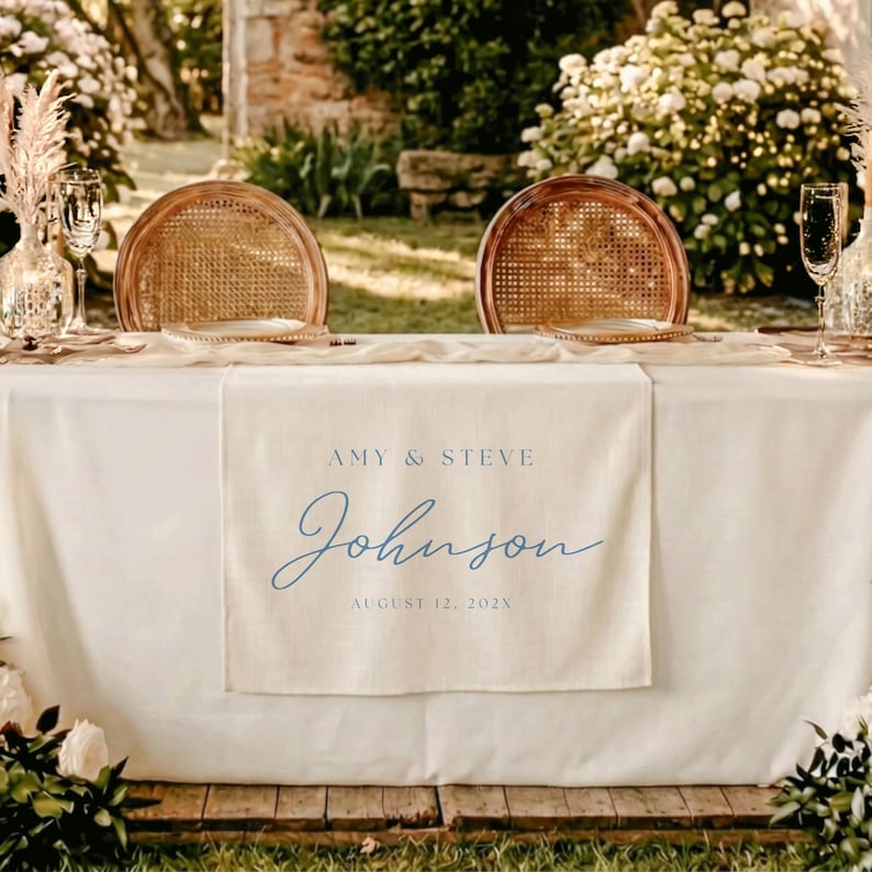 May include: A cream-colored fabric table runner featuring blue printed text that reads AMY & STEVE Johnson AUGUST 12, 202X. The runner is draped over a white tablecloth between two round wooden chairs with woven cane backrests and clear glassware.