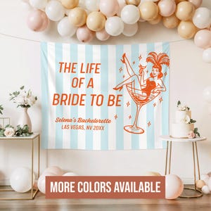 May include: A light blue and white striped backdrop with the text "THE LIFE OF A BRIDE TO BE" in orange. The backdrop features a retro illustration of a woman in a martini glass. The text "Selena's Bachelorette, Las Vegas, NV 20XX" is also visible.
