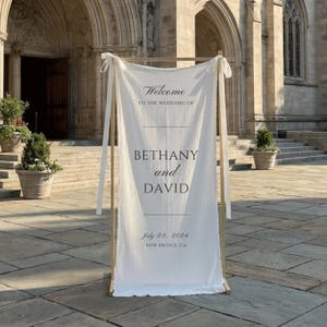Może przedstawiać: Pionowy znak powitalny na ślub z białej tkaniny zawieszony na drewnianej ramie. Tekst brzmi: Welcome TO THE WEDDING OF BETHANY and DAVID July 24, 2026 NEW BRIDGE, CA. Tkanina jest przymocowana w g&oacute;rnych rogach za pomocą białych wstążek.