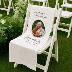 May include: A white fabric memorial sign draped over a white folding chair. The sign features the text RESERVED in loving memory of, a circular photograph of two people embracing, and the name James Sanchez, FATHER OF THE BRIDE, OCT. 13, 20XX.