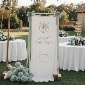 May include: A white fabric sign with the text "Her Next Chapter Begins" and "Emma's Bridal Shower" in elegant script. The sign is decorated with a floral book illustration and sage green ribbons. The scene is set outdoors with round tables.