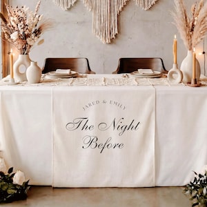May include: A cream-colored fabric table runner draped over a table, featuring the printed black text "JARED & EMILY The Night Before". The table is set with white ceramic vases, taper candles, and plates, with a macrame runner overlay.