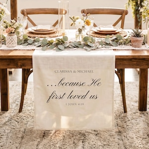 May include: A cream-colored fabric table runner featuring the printed text CLARISSA & MICHAEL, ...because He first loved us, and 1 JOHN 4:19 in black lettering. The runner is draped over a wooden table set with plates, glasses, and greenery.