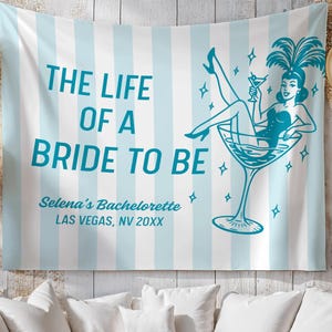 May include: A light blue and white striped tapestry with the text "THE LIFE OF A BRIDE TO BE". It features a teal illustration of a woman in a martini glass. The text "Selena's Bachelorette LAS VEGAS, NV 20XX" is also included.