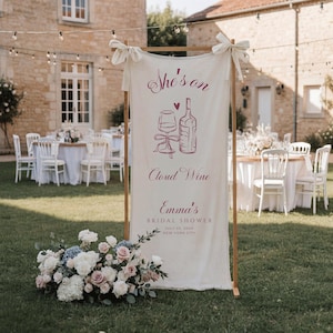 May include: A vertical off-white fabric banner on a wooden frame featuring dark pink text and illustrations of a wine glass and bottle. The text reads: She's on Cloud Wine, Emma's BRIDAL SHOWER, JULY 23, 20XX, NEW YORK CITY. A floral arrangement sits at the base.