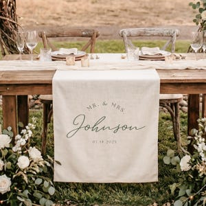 May include: A cream-colored linen table runner draped over a wooden table, featuring the printed text MR. & MRS. Johnson and the date 07.18.202X in a dark green script and serif font, centered on the fabric hanging off the table edge.