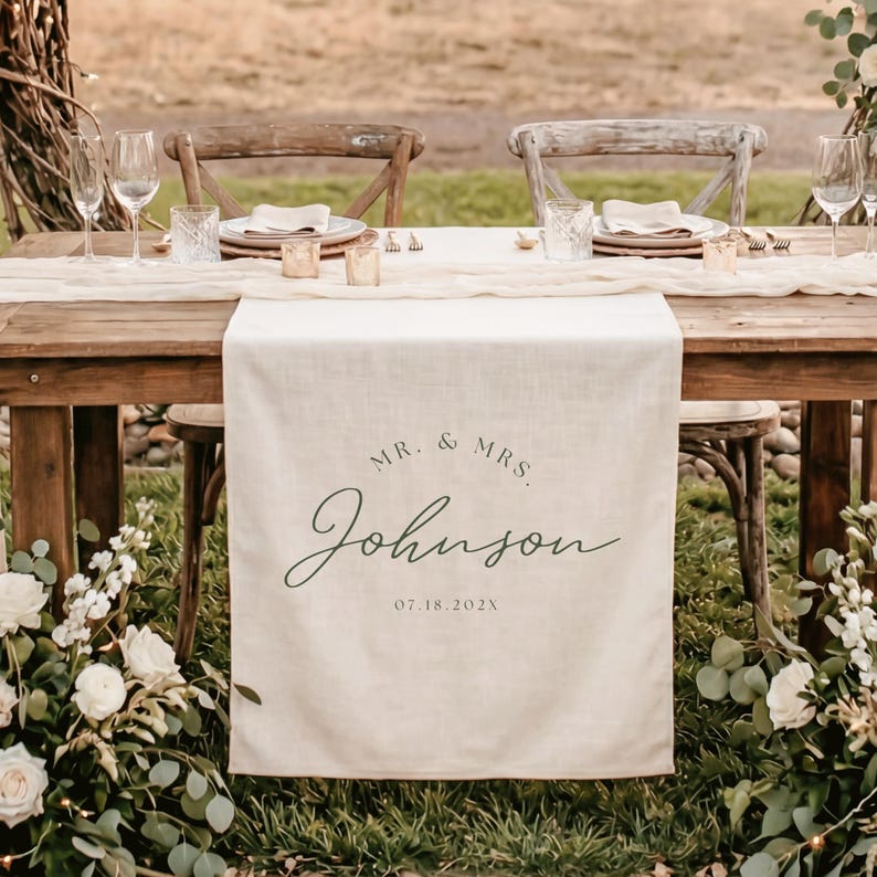 May include: A cream-colored linen table runner draped over a wooden table, featuring the printed text MR. & MRS. Johnson and the date 07.18.202X in a dark green script and serif font, centered on the fabric hanging off the table edge.