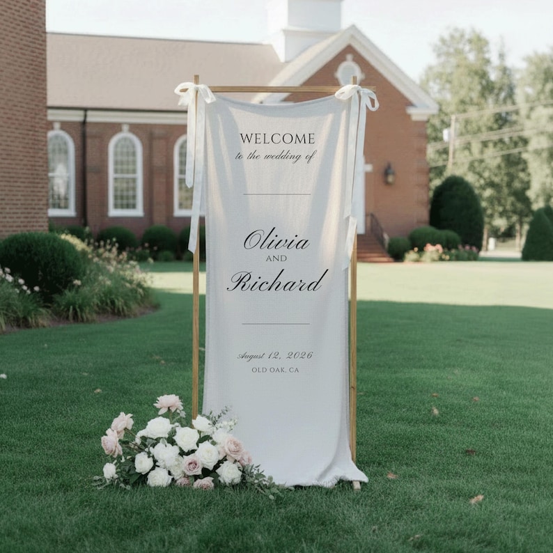 May include: A wedding welcome sign with the text "WELCOME to the wedding of Olivia and Richard" on a white fabric. The sign is framed by a wooden structure and decorated with a floral arrangement. The date August 12, 2026, and location Old Oak, CA are also displayed.