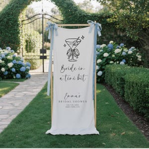May include: A white banner with a wooden frame and light blue ribbons. The banner features a black cocktail illustration and the text "Bride in a tini bit." Below, it reads "Emma's Bridal Shower, July 23, 20xx, New York City."