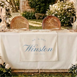 May include: A rectangular cream-colored fabric table runner featuring blue printed text that reads MR. & MRS. Winston EST. 202X. The runner is draped over a white tablecloth, positioned between two round wooden chairs with woven cane backrests.
