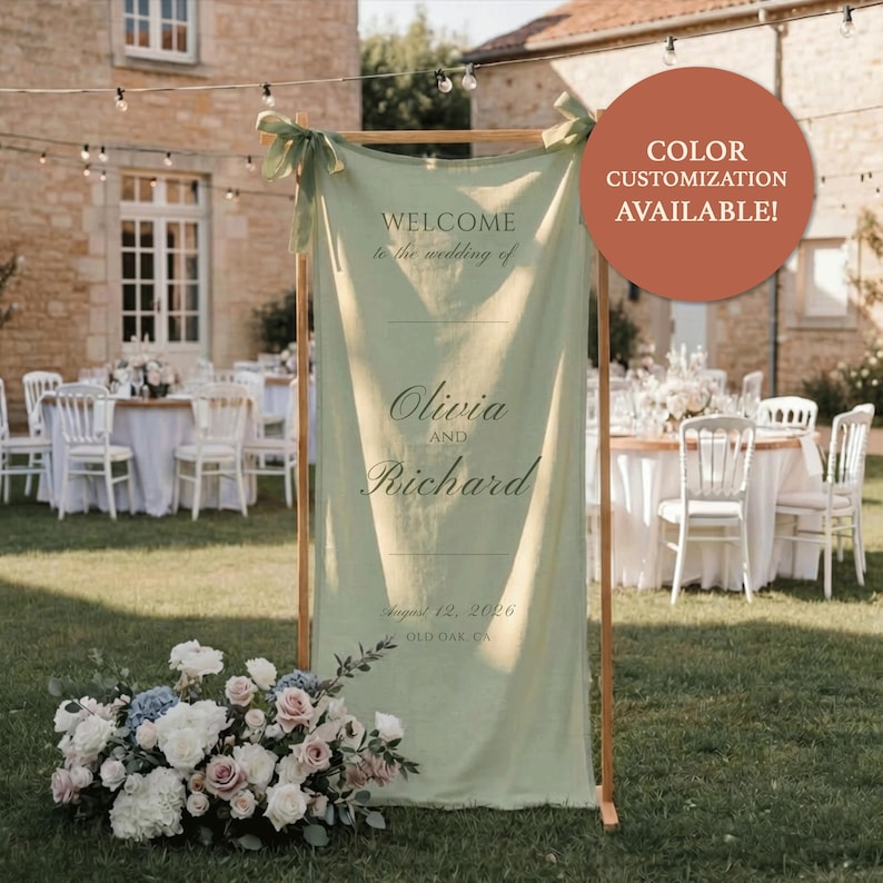 May include: A sage green wedding welcome sign with the text "Welcome to the wedding of Olivia and Richard" and the date August 12, 2026. The sign is displayed on a wooden frame, with a floral arrangement at the base.