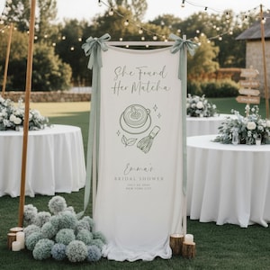 May include: A tall, vertical white fabric banner suspended on a wooden frame with sage green ribbons. The banner features dark green text reading 'She Found Her Matcha', 'Emma's BRIDAL SHOWER', 'JULY 23, 20XX', and 'NEW YORK CITY', with line art of a matcha bowl and whisk.