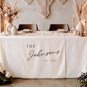 May include: A cream-colored fabric table runner featuring the black printed text THE Johnsons est. 202X centered on the front panel. The runner is draped over a table set with white plates, beige candles in circular holders, and dried floral arrangements.