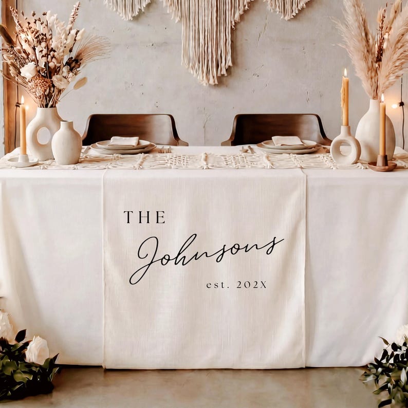 May include: A cream-colored fabric table runner featuring the black printed text THE Johnsons est. 202X centered on the front panel. The runner is draped over a table set with white plates, beige candles in circular holders, and dried floral arrangements.