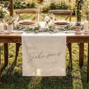 May include: A cream-colored linen table runner featuring the printed text THE Johnsons est. 202X in a brown serif and script font. The runner is draped over a wooden table set with eucalyptus garlands, glass candle holders, and floral arrangements.