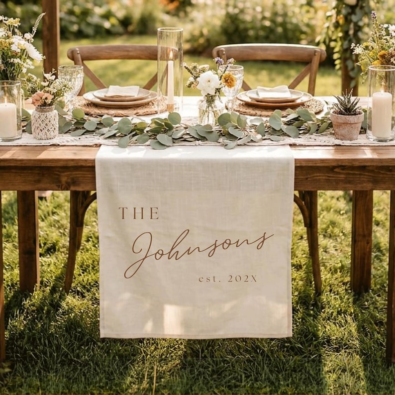 May include: A cream-colored linen table runner featuring the printed text THE Johnsons est. 202X in a brown serif and script font. The runner is draped over a wooden table set with eucalyptus garlands, glass candle holders, and floral arrangements.