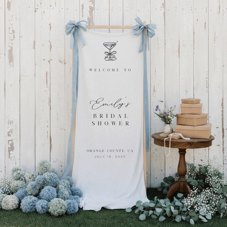 Puede incluir: Una pancarta de tela blanca con una ilustraci&oacute;n de c&oacute;ctel y el texto "WELCOME TO Emily's BRIDAL SHOWER". La pancarta est&aacute; suspendida de un marco de madera con cintas azul claro. Una peque&ntilde;a mesa contiene regalos y flores.