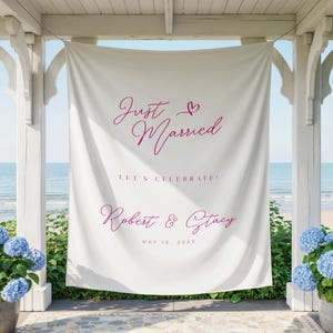 May include: A white fabric banner with the words "Just Married" in a cursive font, along with a heart symbol. Below, it reads "LET'S CELEBRATE!" and the names "Robert & Stacy" with a date. The banner is displayed outdoors.