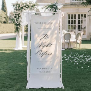 May include: A white vertical fabric banner suspended on a white wooden frame, featuring the text "TYLER & JULIANNE", "The Night Before", "NEW BRIDGE, CA", and "JULY 18, 202X" in black lettering, with white ribbons tied to the top corners of the frame.