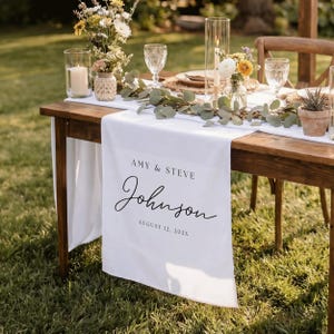 May include: A white fabric table runner draped over a wooden table, featuring the printed text AMY & STEVE Johnson AUGUST 12, 202X. The runner is decorated with a green eucalyptus garland, floral arrangements, glass candle holders, and clear glasses.