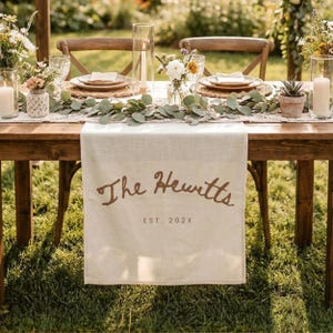 May include: A cream-colored linen table runner draped over a wooden table, featuring the brown printed text The Hewitts and EST. 202X. The table is decorated with eucalyptus garlands, glass candle holders with white candles, and floral arrangements.