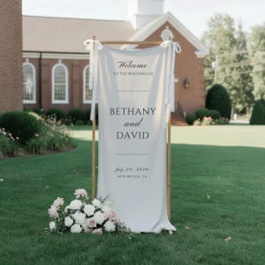 Może przedstawiać: Wysoki, kremowy szyld powitalny z tkaniny zawieszony na drewnianej ramie, z czarnym napisem: Welcome TO THE WEDDING OF BETHANY and DAVID July 24, 2026 NEW BRIDGE, CA. U podstawy znajduje się kompozycja kwiatowa z białych i r&oacute;żowych r&oacute;ż.