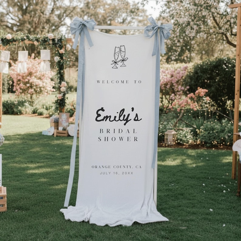 Puede incluir: Una pancarta de tela blanca con cintas azul claro, que da la bienvenida a los invitados a "Emily's Bridal Shower". La pancarta presenta copas de champ&aacute;n y detalles del evento: Orange County, CA, 16 de julio de 20XX. El letrero se muestra al aire libre en un c&eacute;sped verde.