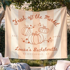 Może przedstawiać: Beżowo-biała, pasiasta gobelina z napisem "Fresh off the Market" i ilustracjami dyni. Gobelin zawiera również tekst "Louisa's Bachelorette" i "Scottsdale, AZ".