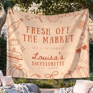 Może przedstawiać: Beżowy baner z tkaniny z brązowym napisem "FRESH OFF THE MARKET". Baner zawiera ilustracje dyni, butelki wina, kosza i kieliszków do wina. Wyświetlany jest również tekst "Louisa's Bachelorette".