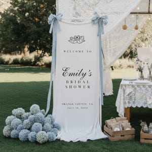 Puede incluir: Una pancarta blanca con el texto "Welcome to Emily's Bridal Shower" y una ilustraci&oacute;n de cisnes. La pancarta est&aacute; decorada con cintas azul claro y est&aacute; colocada al aire libre sobre un c&eacute;sped verde. La fecha y el lugar tambi&eacute;n est&aacute;n impresos en la pancarta.