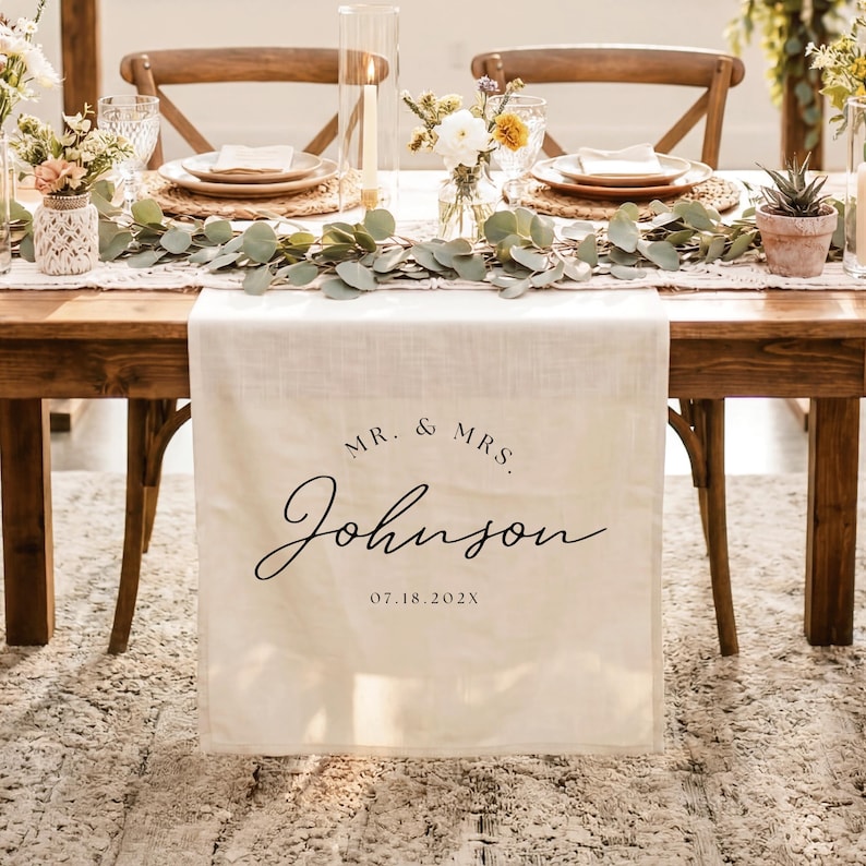 May include: A cream-colored fabric table runner featuring the black printed text MR. & MRS. Johnson and the date 07.18.202X. The runner hangs over the edge of a wooden table decorated with eucalyptus greenery, floral arrangements, and candles.