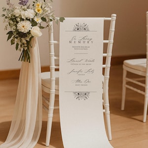 May include: A white chair with a floral arrangement and a cream-colored banner. The banner reads "In Loving Memory" with names and titles below. The chair is decorated for a memorial or wedding.