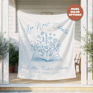 Puede incluir: Bandera de tela blanca con texto azul e ilustraciones florales. La bandera dice "Her Next Chapter" sobre un libro abierto con flores y el texto "Libby's Bridal Shower, New York 20XX". Un círculo marrón con "MORE COLOR OPTIONS" está en la parte superior derecha.