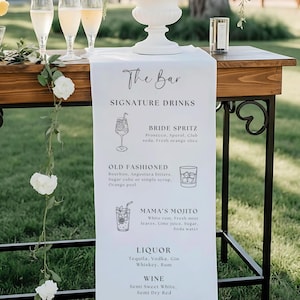 May include: A white fabric bar menu runner featuring black text and line art illustrations of cocktails. The menu lists The Bar, Signature Drinks including Bride Spritz, Old Fashioned, and Mama's Mojito, followed by sections for Liquor and Wine.
