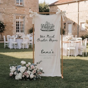 May include: A vertical off-white fabric banner suspended on a wooden frame, featuring a black line drawing of flowers in an open book, with the text "Her Next Chapter Begins", "Emma's BRIDAL SHOWER", "JULY 23, 20XX", and "NEW YORK CITY".