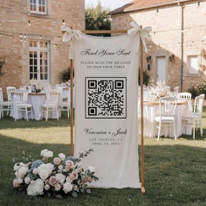 May include: A vertical cream-colored fabric sign suspended on a wooden frame, featuring the text "Find Your Seat," "PLEASE SCAN THE QR CODE TO FIND YOUR TABLE," "Veronica + Jack," "LOS ANGELES, CA," and "APRIL 24, 20XX" with a large black QR code.
