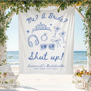 May include: White fabric banner with blue text and illustrations for a bachelorette party. The banner reads "Me? A Bride? Shut up!" with illustrations of a tiara, headphones, ring, sunglasses, bow, and martini glasses. The banner also includes "Savannah's Bachelorette NEW YORK CITY 2025".