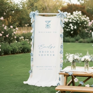 Puede incluir: Un letrero de tela blanca con detalles azules y el texto "Bienvenida a la despedida de soltera de Emily". El letrero est&aacute; decorado con lazos azules y detalles florales. Una mesa de madera con arreglos florales est&aacute; frente al letrero.