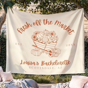 Może przedstawiać: Kremowy baner z napisem "Fresh off the Market" w kolorze rdzy. W centrum znajduje się taczka wypełniona dyniami i kwiatami. Poniżej napis "Louisa's Bachelorette, Scottsdale, AZ".