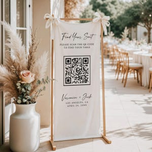May include: A white fabric wedding seating chart sign suspended from a wooden frame with cream ribbons. The sign features black text reading Find Your Seat, Please Scan The QR Code To Find Your Table, Veronica + Jack, Los Angeles, CA, April 24, 20XX.