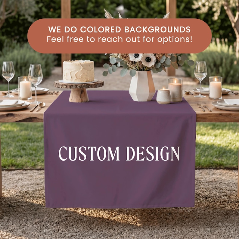 May include: A solid dark purple table runner featuring the white text CUSTOM DESIGN centered on the front. Above the runner, a white cake on a wooden stand, a white vase with flowers, and two glass candles are arranged on a wooden table.