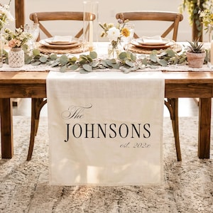 May include: A cream-colored fabric table runner featuring the black printed text The JOHNSONS est. 202x, draped over a wooden table. The runner displays a personalized family name and year in a combination of serif and elegant script typography.