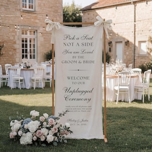 May include: A tall, rectangular off-white fabric sign suspended on a wooden frame, featuring black printed text that reads: Pick a Seat NOT A SIDE You are Loved BY THE GROOM & BRIDE WELCOME TO OUR Unplugged Ceremony, with names Hailey & Justin.